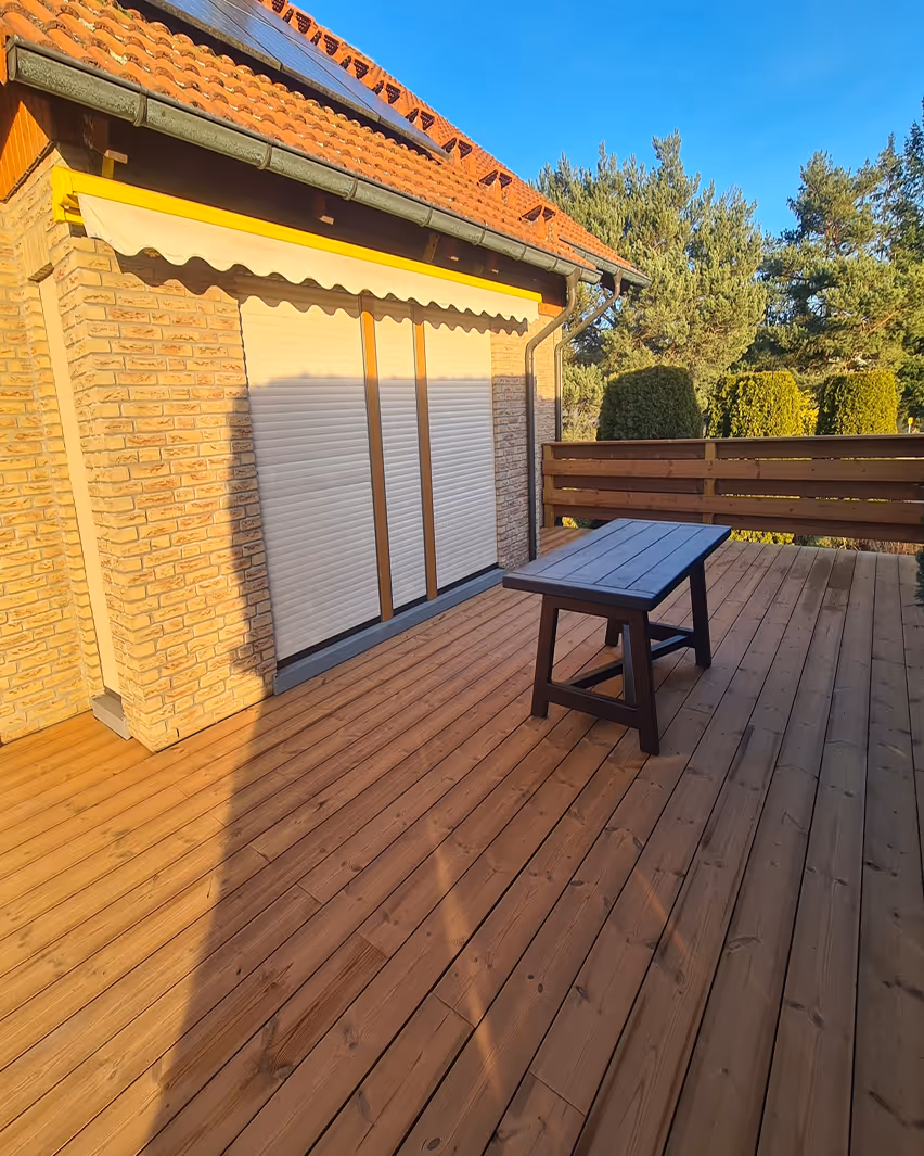 Holzterrasse mit einem Tisch vor einem Haus mit gelber Markise und geschlossenen Rollläden, umgeben von Bäumen.
