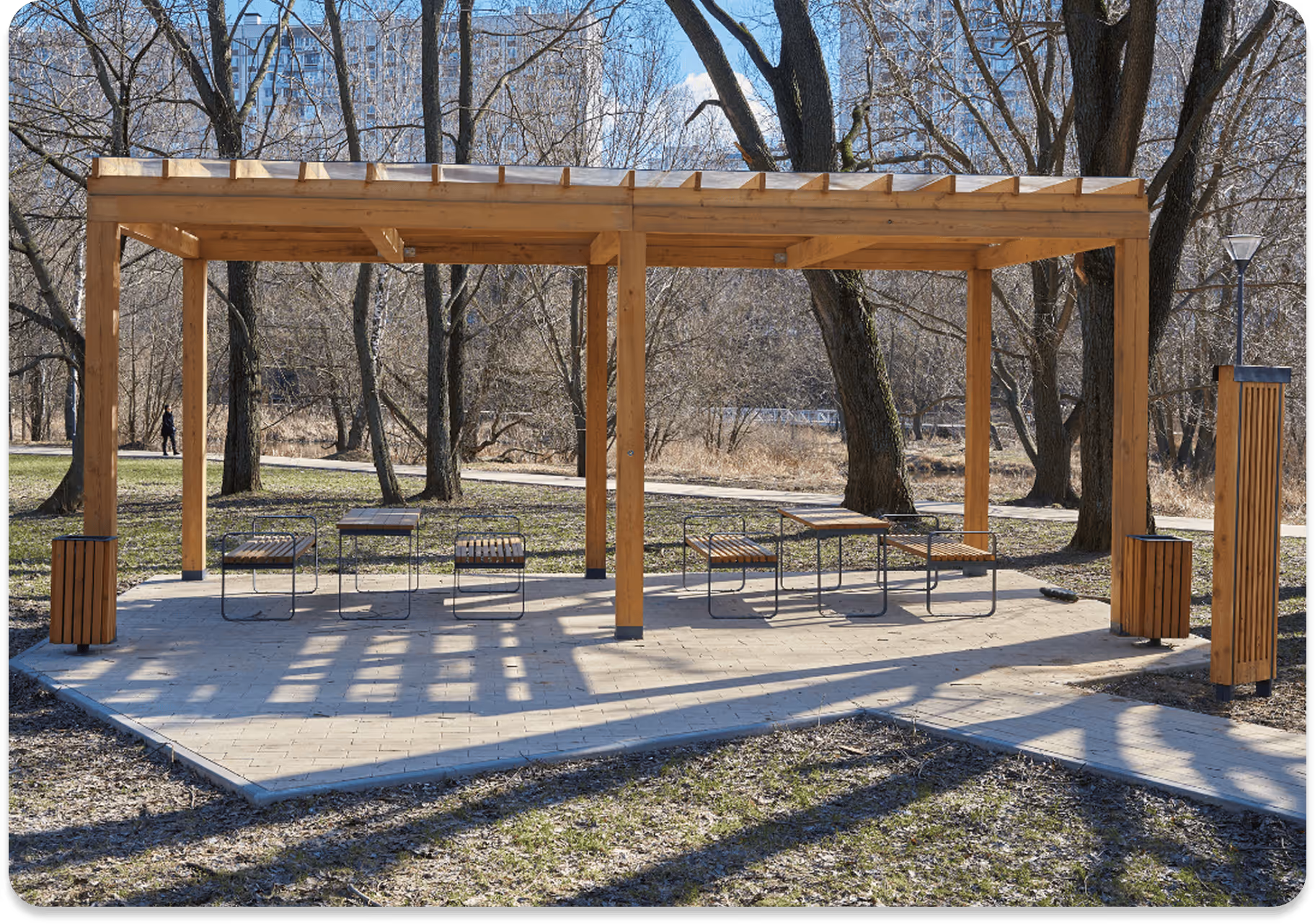 Moderner Pavillon aus Holz mit Sitzbänken und Tischen in einem Park an einem sonnigen Tag.