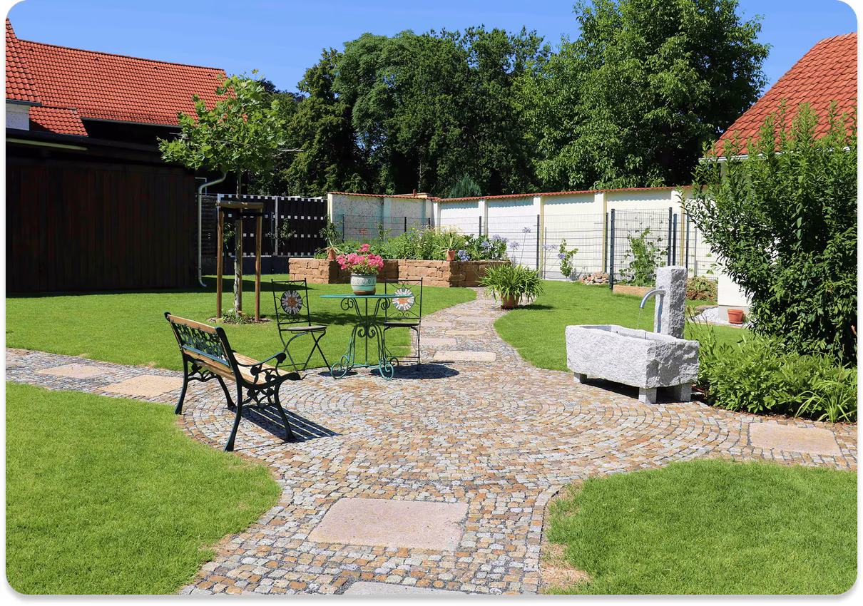 Gepflegter Garten mit gepflastertem Weg, Bänken, Tisch mit zwei Stühlen, Blumen und Steinbrunnen.