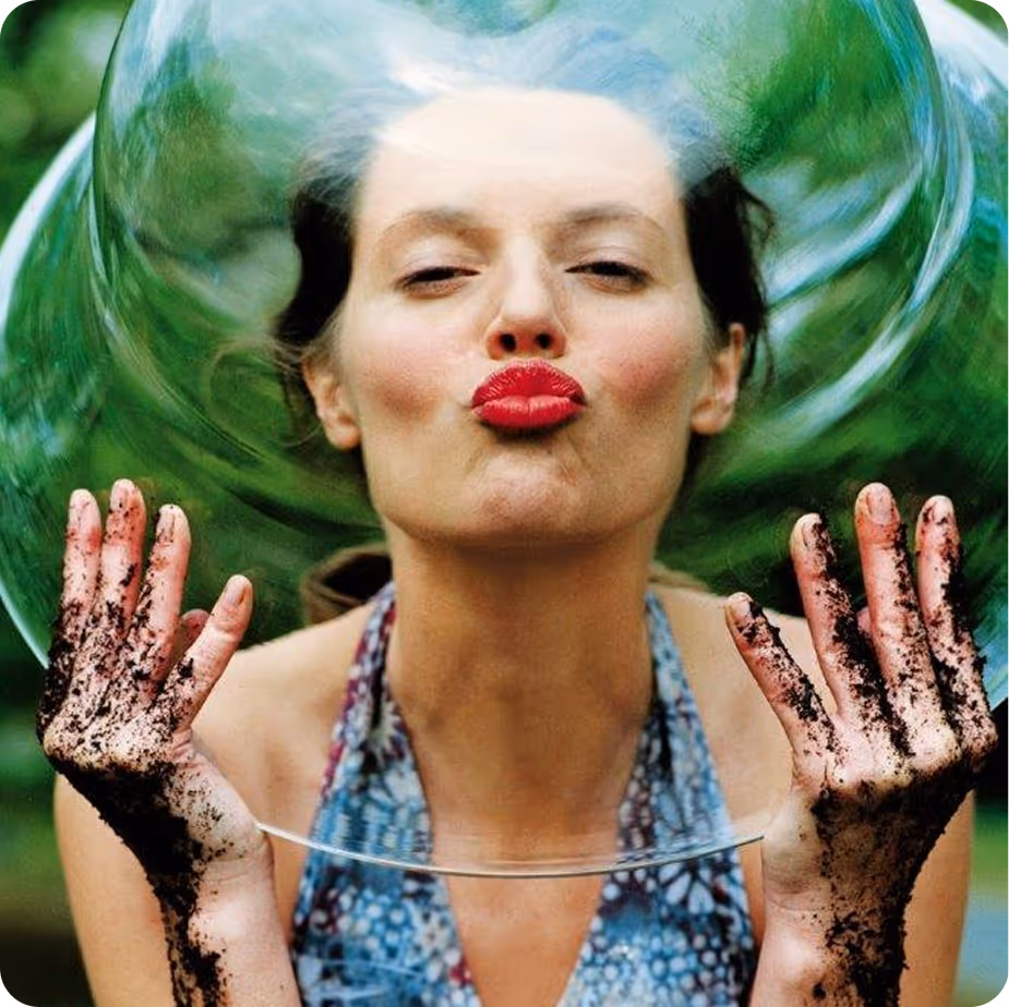 Frau mit roten Lippen, die ein großes, durchsichtbares Seifenblasen um ihr Gesicht hält, ihre Hände sind mit Erde bedeckt.