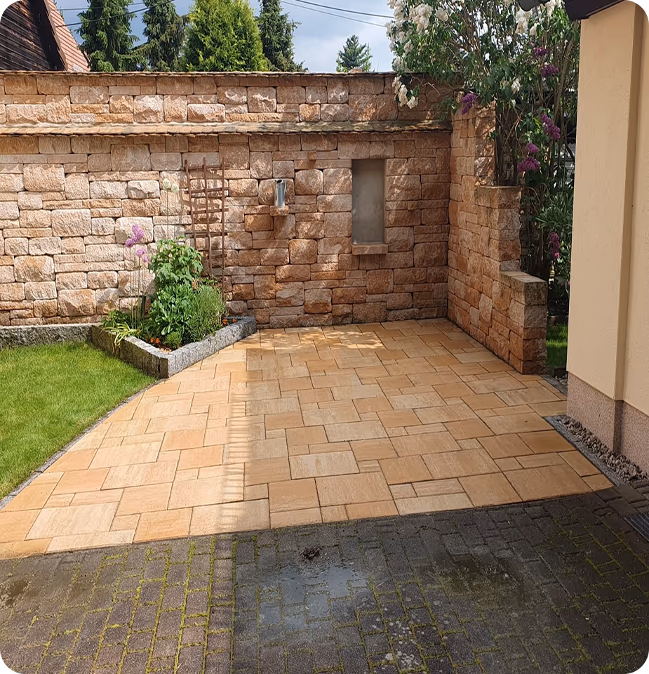 Gepflasterte Terrasse mit einer niedrigen Steinmauer, einem kleinen Blumenbeet und Rasen unter blühenden Sträuchern.
