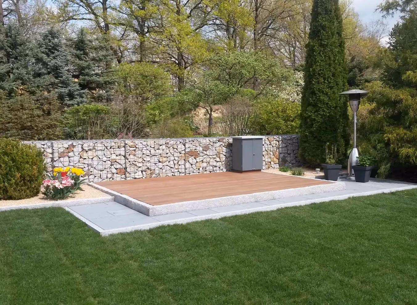 Moderner Garten mit einer Holzplattform vor einer Gabionensteinmauer, umgeben von Blumen, grünen Sträuchern und Bäumen.