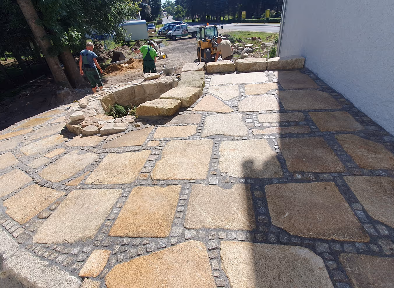 Handwerker bauen eine Terrasse mit großen Steinplatten, im Hintergrund Bagger und Fahrzeuge.
